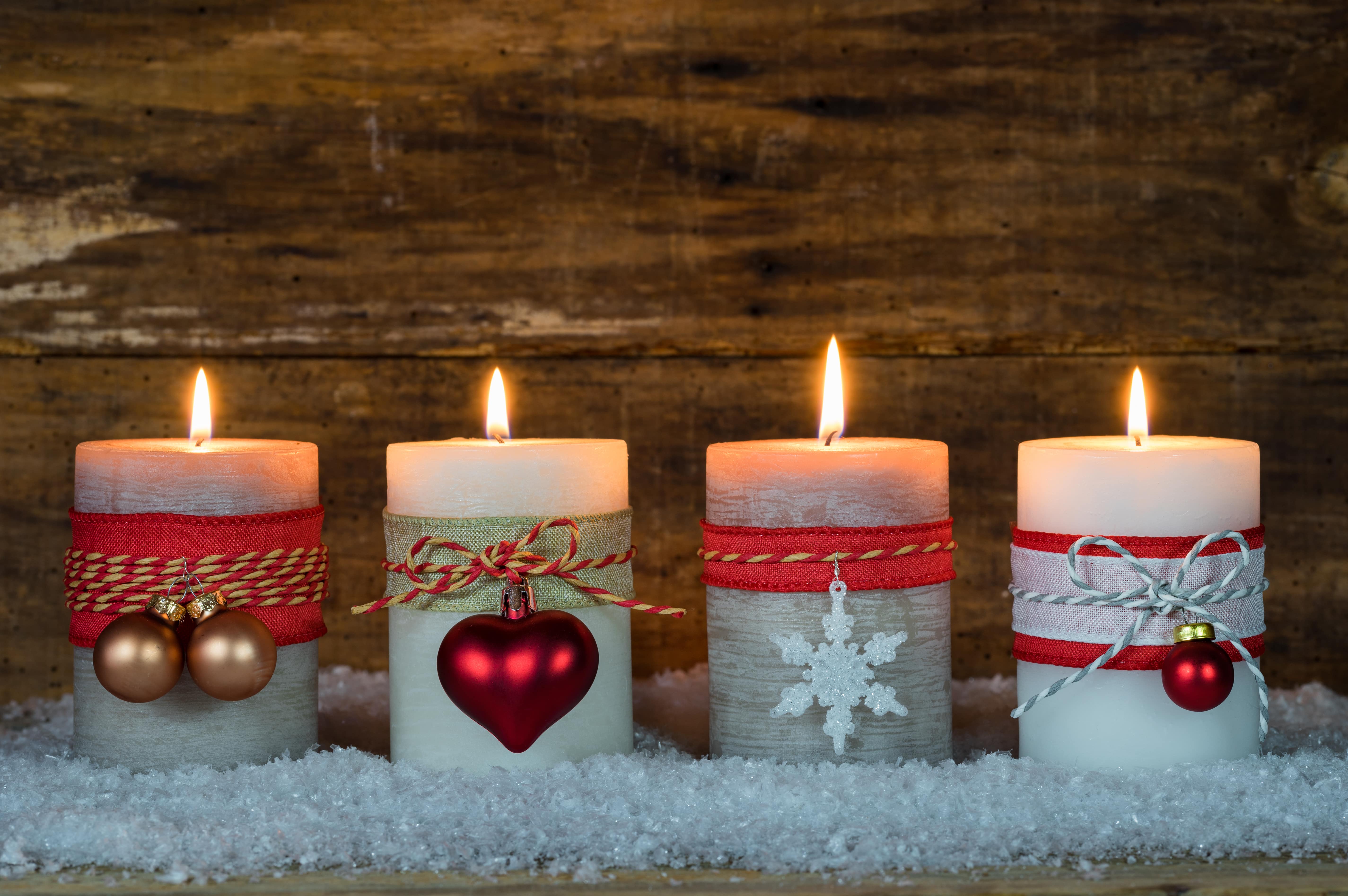 Four lit candles wrapped with ribbon and ornaments sitting on top of artificial snow in front of a wooden backdrop